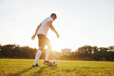 Arka planda orman var. Genç futbolcu spor sahasında antrenman yapıyor..