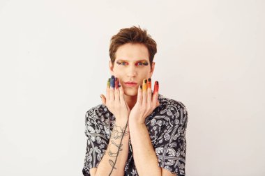 Genç eşcinsel adam stüdyoda duruyor ve kameraya poz veriyor. Yüz parmaklarında çok renkli makyaj malzemeleri var..