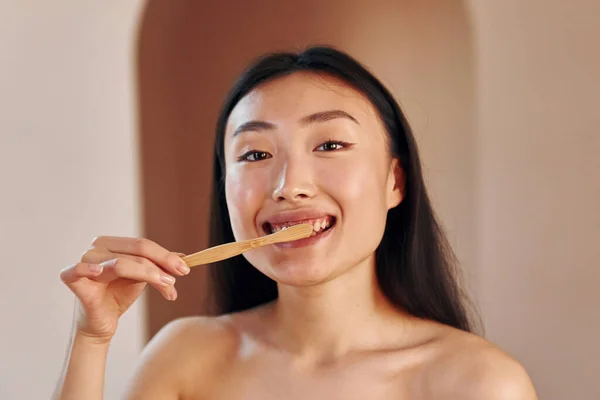Holds toothbrush. Young serious asian woman standing indoors.