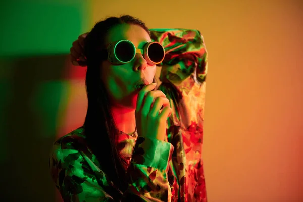 In retro pilot glasses. Fashionable young woman standing in the studio with neon light.