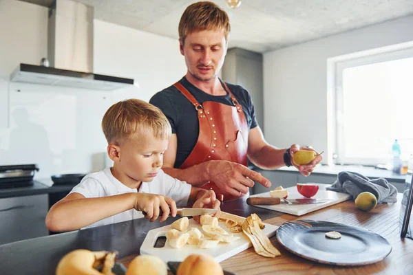 Man is in the apron on kitchen. Father and son is indoors at home together.
