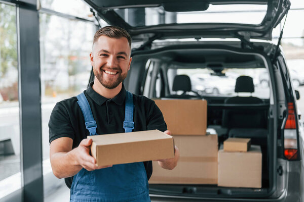 Holding box. Delivery man in uniform is indoors with car and with order.