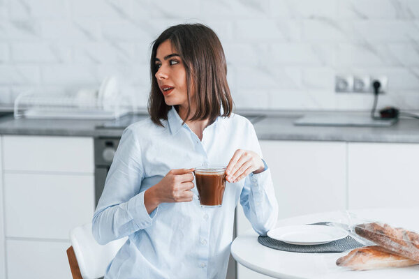 With cup of coffee. Young woman is indoors in room of modern house at daytime.