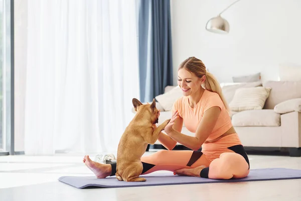 On fitness mat. Woman with pug dog is at home at daytime.