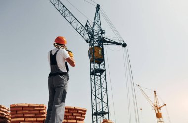 Arka planda vinç var. Üniformalı genç inşaat işçisi tamamlanmamış binada meşgul..