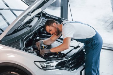 Kaput açıldı. Beyaz gömlekli ve mavi üniformalı genç adam otomobil tamir ediyor..