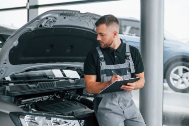 Not defteriyle. Üniformalı adam gündüz vakti otosalonda çalışıyor..