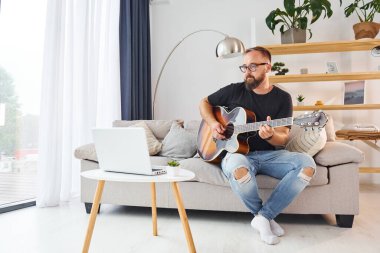 Laptop kullanıyorum. Gündelik giysiler içinde ve akustik gitarlı bir adam içeride..
