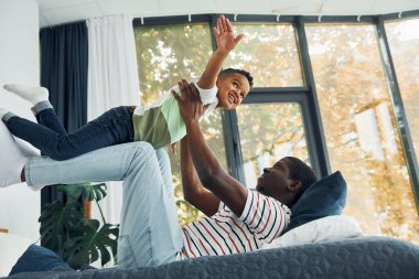 Yatağa uzanıp eğlenmek. Afrikalı Amerikalı baba ve küçük oğlu evde..