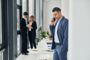 Adam telefonla konuşuyor. Ofiste resmi giysiler içinde bir grup insan var..