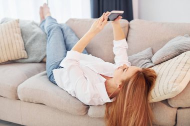 Ellerinde telefonla. Beyaz gömlekli ve kot pantolonlu genç bayan evde..