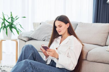 Ellerinde telefonla kanepenin yanında oturuyor. Beyaz gömlekli ve kot pantolonlu genç bayan evde..