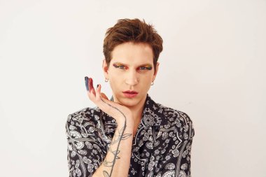 Genç eşcinsel adam stüdyoda duruyor ve kameraya poz veriyor. Yüz parmaklarında çok renkli makyaj malzemeleri var..