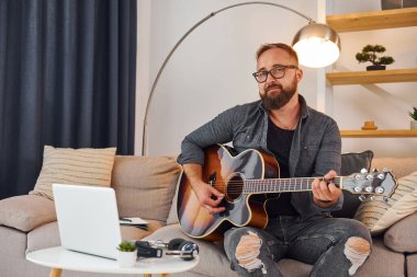 Kanepede. Akustik gitar çalıyor. Sıradan elbiseli adam içeride..