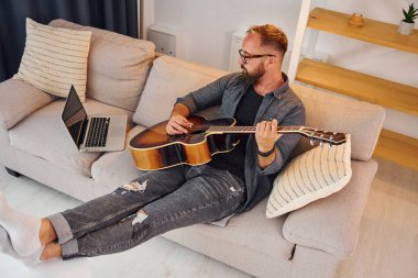 Kanepede. Akustik gitar çalıyor. Sıradan elbiseli adam içeride..
