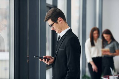 Telefonu olan genç bir adam. Ofiste resmi giysiler içinde bir grup insan var..