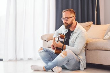 Kanepenin yanında oturuyor. Gündelik giysiler içinde ve akustik gitarlı bir adam içeride..