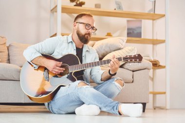 Kanepenin yanında oturuyor. Gündelik giysiler içinde ve akustik gitarlı bir adam içeride..