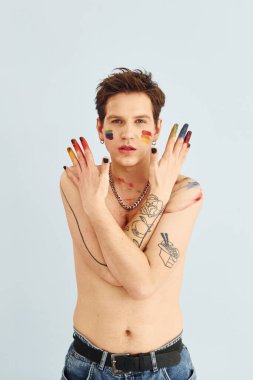 Genç eşcinsel adam stüdyoda duruyor ve kameraya poz veriyor. Gökkuşağı makyajlı tişörtsüz adam..