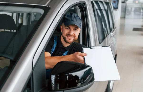 Sitting in vehicle and holding document. Delivery man in uniform is indoors with car and with order.