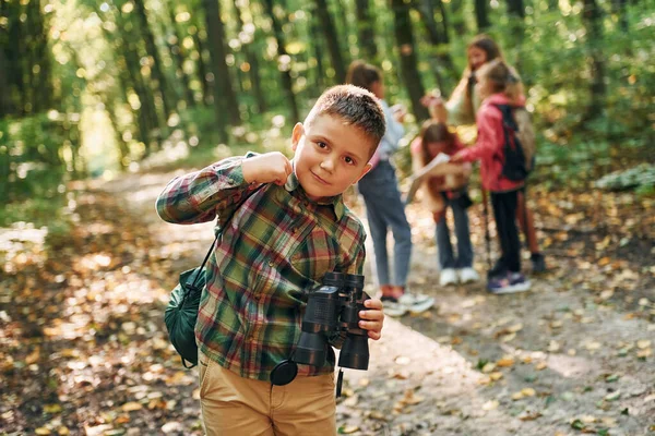 一个拿着双筒望远镜站在朋友面前的男孩 夏天的白天 孩子们一起在绿林里 — 图库照片