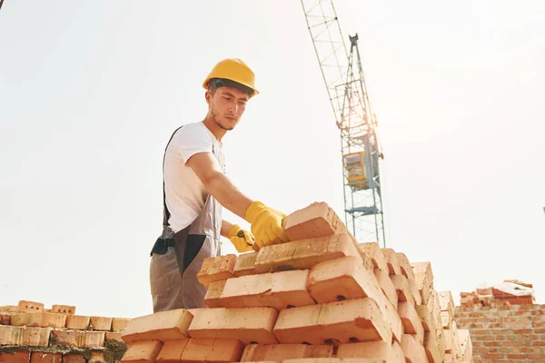 Using bricks. Young construction worker in uniform is busy at the unfinished building.