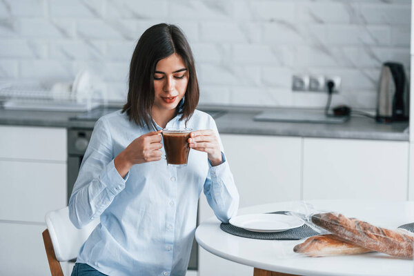 With cup of coffee. Young woman is indoors in room of modern house at daytime.