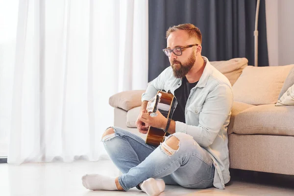 Sitting near the sofa. Man in casual clothes and with acoustic guitar is indoors.