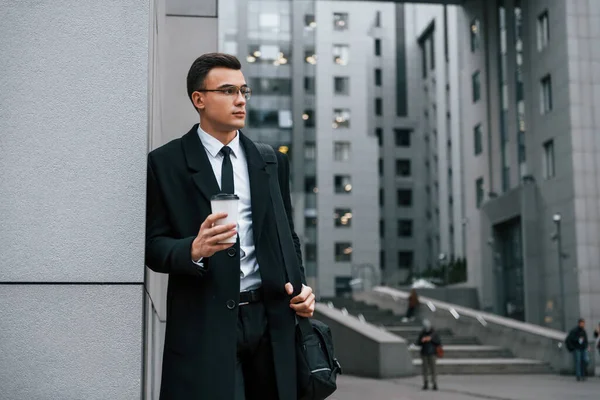 Standing near the building. Businessman in black suit and tie is outdoors in the city.