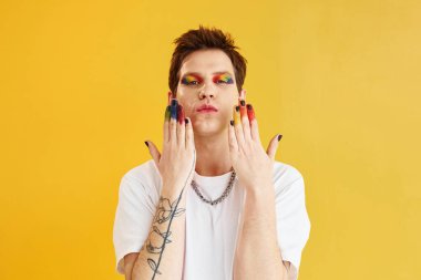 Genç eşcinsel adam stüdyoda duruyor ve kameraya poz veriyor. Gökkuşağı makyajı.