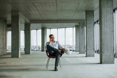 Telefonla konuşuyor. Resmi kıyafetli genç adam içeride inşaatta çalışıyor..