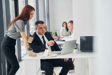 Dizüstü bilgisayarla çalışıyor. Ofiste resmi giysiler içinde bir grup insan var..