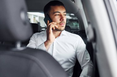 Telefonla konuşuyor. Beyaz gömlekli genç adam modern bir otomobilin içinde oturuyor..