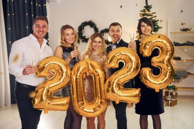 2023 numaralı balonları tutuyorum. Bir grup insan içeride yeni yıl partisi veriyor..
