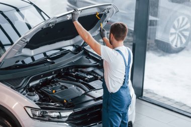 Kaput açıldı. Beyaz gömlekli ve mavi üniformalı genç adam otomobil tamir ediyor..