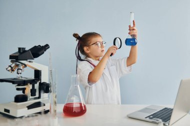 Büyüteç tutuyor. Palto giyen küçük bir kız laboratuarda ekipman kullanarak bilim adamını oynuyor..