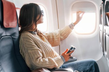 Sitting with phone in hands. Young female passanger in casual clothes is in the plane. clipart