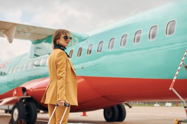 Sarı elbiseli, güneş gözlüklü ve bagajlı yolcu uçağın yanında..