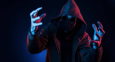 Gözlüklü, maskeli ve başlıklı korkunç adam. Neon ışıkları. Genç Avrupalı adam karanlık stüdyoda..