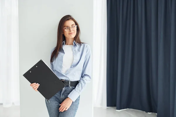 Young woman in glasses, formal clothes and with notepad standing indoors.
