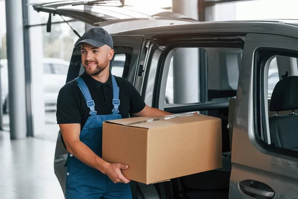 With box in hands. Delivery man in uniform is indoors with car and with order.