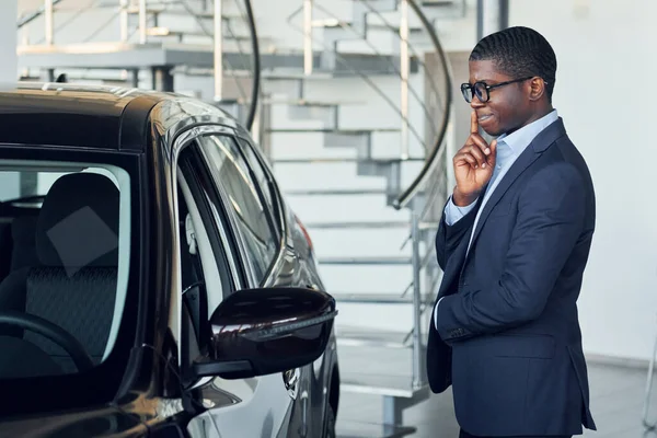 Standing near car. Young african american businessman in black suit is the autosalon.