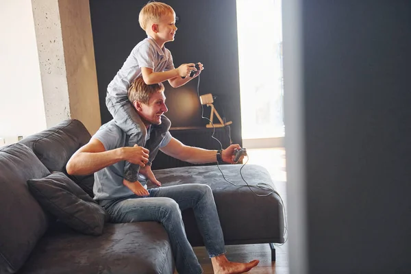 Boy is sitting on the man\'s shoulders. Father and son is indoors at home together.