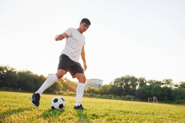 Arka planda orman var. Genç futbolcu spor sahasında antrenman yapıyor..
