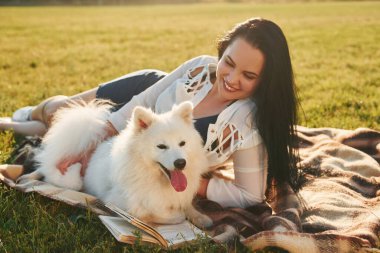 Kitapla. Köpeğiyle bir kadın güneşli bir günde sahada eğleniyor..