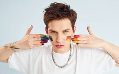 Genç eşcinsel adam stüdyoda duruyor ve kameraya poz veriyor. Çok renkli makyajla boyanmış..