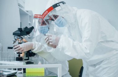 Mikroskop kullanarak. Koruyucu üniformalı kadın tıp çalışanları COVID-19 aşısı üzerinde çalışıyorlar..