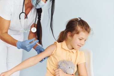 Oyuncak ayıyla küçük bir kız. Üniformalı doktor hastaya aşı yapıyor..