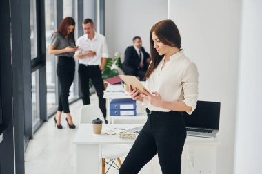Kadının elinde tablet var. Ofiste resmi giysiler içinde bir grup insan var..