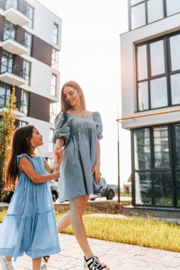 Güneş ışığıyla aydınlatılmış. Küçük kızıyla birlikte binaların etrafında yürüyen genç bir anne..